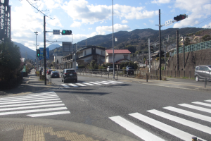 Eine Straße in der Stadt mit Autos, einer Zebürürderung, Strommasten, Verkehrsampeln, Gebäuden, Bäumen, Höhen und einem bewölktem Himmel.