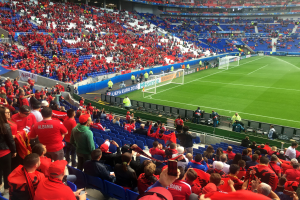 Ein volles Stadion mit Sitz- und Stehplätzen, Bannern, einem Tor und Deckenleuchten, während Fans gespannt auf ein Fußballspiel warten.
