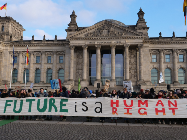 Eine Gruppe von Menschen hält ein Transparent mit der Aufschrift 'Zukunft ist ein Menschenrecht' vor dem Reichstagsgebäude in Berlin, Deutschland, mit sichtbaren Fenstern, Säulen und Bögen, umgeben von Fahnenmasten und einem bewölkten Himmel.