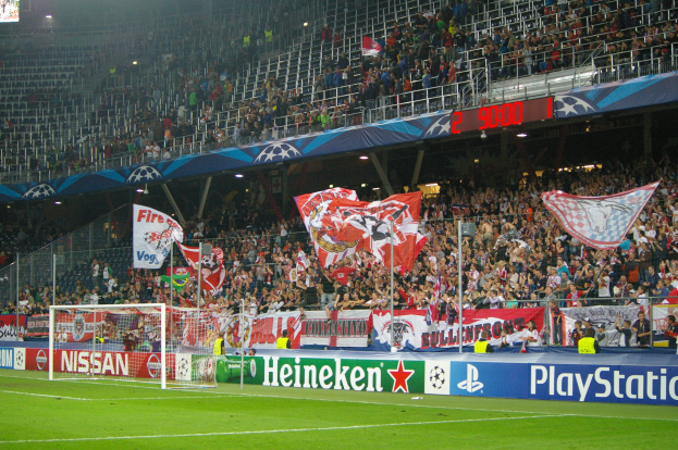 Ein Fussballspiel wird in einem hell erleuchteten Stadion mit einer grossen Zuschauermenge, grünem Rasen, einem Tor mit Bannern und einem Bildschirm in der oberen linken Ecke gespielt.