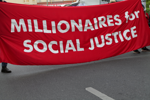 Eine Gruppe von Menschen geht eine Straße entlang und hält eine rote Fahne mit der Aufschrift "Millionäre für soziale Gerechtigkeit" hoch, im Hintergrund sind Gebäude, Pfosten und Bäume zu sehen, unter einem klaren blauen Himmel.