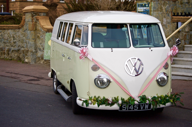 Ein wei├čer und rosa Volkswagen Camper Van, der mit Blumen und Bl├Ąttern geschmückt ist und vor einer Steinwand mit einer Treppe auf der rechten Seite und einem Geb├Ąude im Hintergrund geparkt ist.
