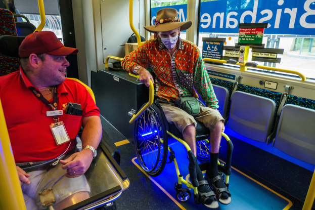 Zwei Männer im Rollstuhl in einem Bus, einer trägt ein rotes T-Shirt und eine Kappe, der andere ein orangefarbenes Hemd und einen braunen Hut, beide tragen Masken und Ausweise, mit gelben Stangen und Glasfenstern, durch die Bäume, Gebäude und der Himmel zu sehen sind.