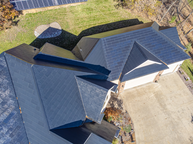 Luftaufnahme eines Hauses mit integrierten Solarpanelen auf dem Dach, umgeben von Bäumen, Gras und einem Zaun.