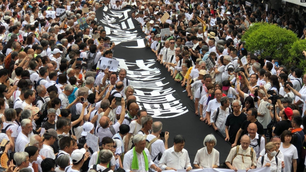 Eine große Gruppe von Menschen marschiert auf der Straße und hält Protestschilder und Banner in der Hand, mit Pflanzen auf der rechten Seite und einem Gebäude im Hintergrund.
