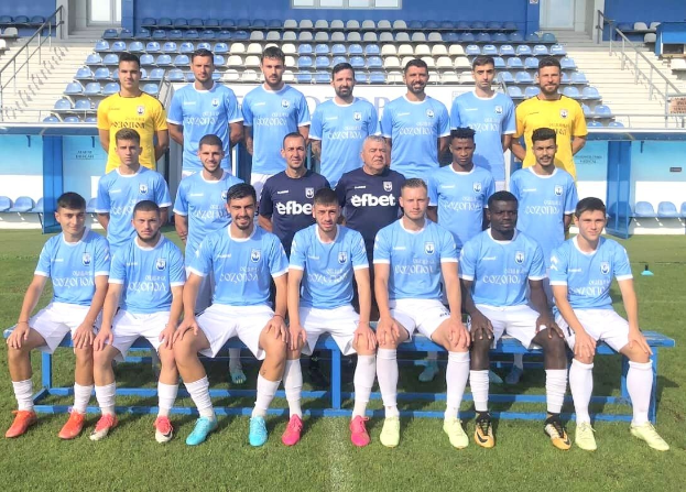 Gruppe von Männern auf einem Fußballfeld, einige sitzen auf Bänken und andere stehen auf dem Rasen, mit Tribünen und architektonischen Elementen im Hintergrund.