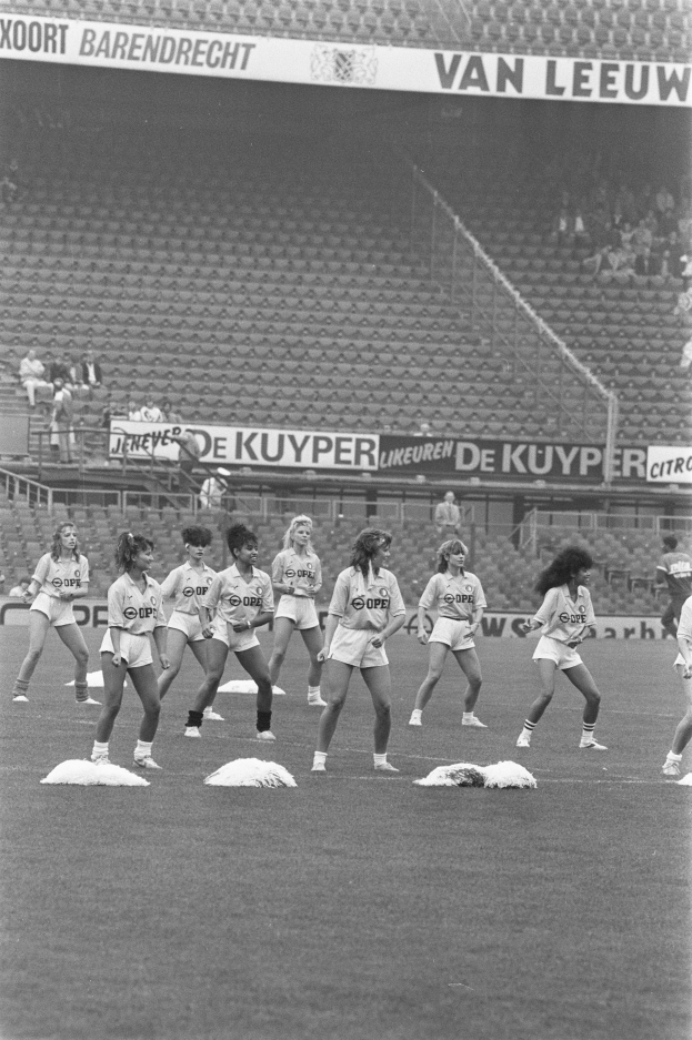 Eine Gruppe von Frauen auf einem Fußballfeld, umgeben von einer Menge im Publikum und Schildern mit Text, auf einem Schwarz-Weiß-Bild.