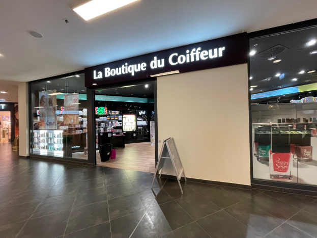 Ein Friseursalon in einem Einkaufszentrum mit einer Texttafel, einem Mülleimer, Regalen mit verschiedenen Gegenständen und einer Decke, die mit Lampen geschmückt ist und so eine einladende Atmosphäre schafft.