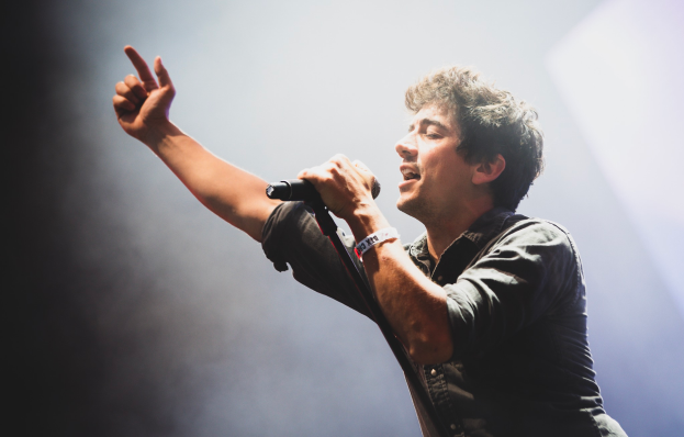 Ein Mann in schwarzem Hemd und Jeans singt leidenschaftlich in ein Mikrofon auf einer Bühne, die Augen geschlossen und das Haar wild gestylt, während das Publikum im Hintergrund jubelt und klatscht.