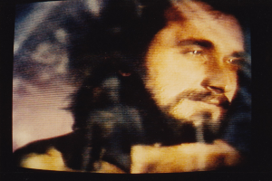 Ein Mann mit Bart und Schnurrbart, langen dunklen Haaren und einem ernsten Gesichtsausdruck, der eine weiße Hemd trägt, schaut direkt in die Kamera auf einem Fernsehbildschirm mit dunklem Hintergrund.