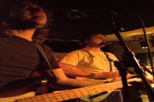 Eine Person in einem wei├čen Hemd singt in ein Mikrofon und spielt eine grüne Gitarre, steht neben einer anderen Person in einem schwarzen Hemd und Brille, die ebenfalls singt und eine schwarze Gitarre spielt, mit Scheinwerfern und anderen Instrumenten im Hintergrund.