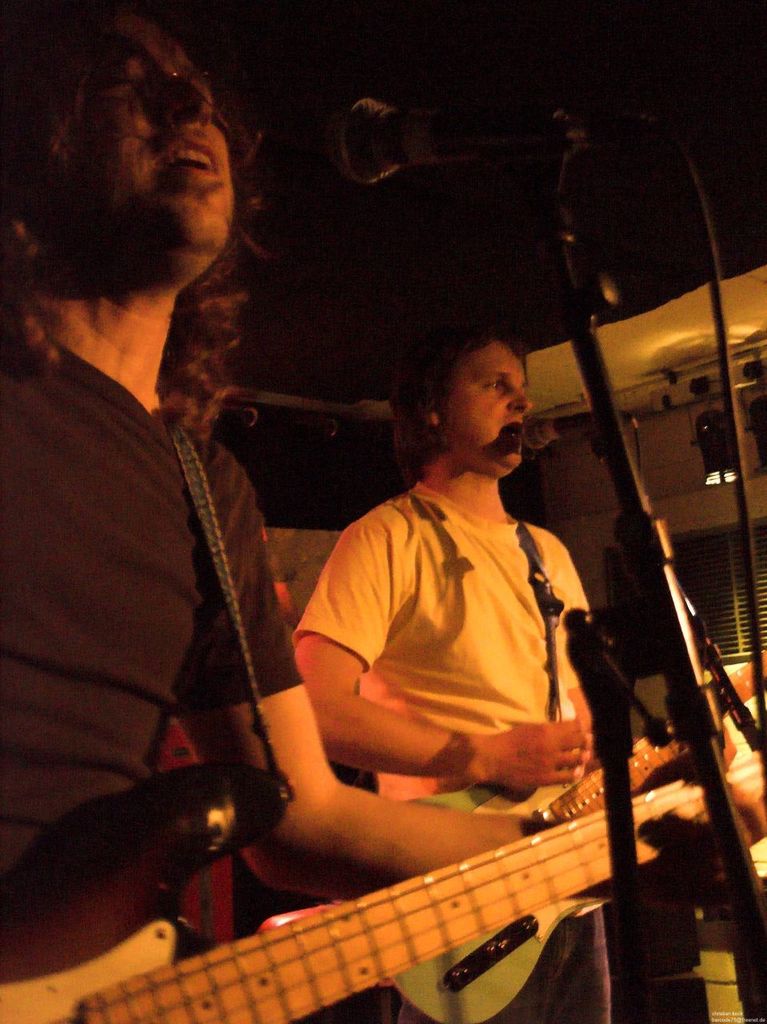 Eine Person in einem wei├čen Hemd singt in ein Mikrofon und spielt eine grüne Gitarre, steht neben einer anderen Person in einem schwarzen Hemd und Brille, die ebenfalls singt und eine schwarze Gitarre spielt, mit Scheinwerfern und anderen Instrumenten im Hintergrund.