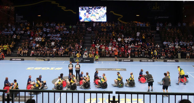 Eine Gruppe von Rollstuhlfahrern auf einem Court bei den Invictus Games Düsseldorf 2020, mit einer großen Zuschauermenge, umgeben von Geländern, Treppen, die zu einem Bildschirm führen, und Informationsschildern.