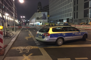 Ein Polizeiauto steht nachts an der Seite einer Straße in Berlin, Deutschland, umgeben von Gebäuden, Straßenlaternen, Straßenschildern, Kraftfahrzeugen, Absperrpoller, Fahrrädern, Menschen auf dem Gehweg, Werbetafeln und einer Statue im Hintergrund, wahrscheinlich nach einem Schusswechsel und einem Autounfall.