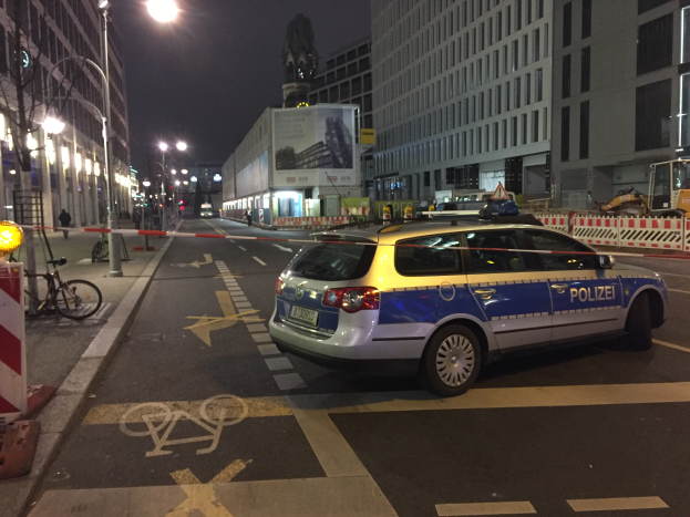 Ein Polizeiauto steht nachts an der Seite einer Straße in Berlin, Deutschland, umgeben von Gebäuden, Straßenlaternen, Straßenschildern, Kraftfahrzeugen, Absperrpoller, Fahrrädern, Menschen auf dem Gehweg, Werbetafeln und einer Statue im Hintergrund, wahrscheinlich nach einem Schusswechsel und einem Autounfall.