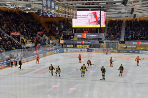 Eine Gruppe von Menschen mit Helmen und Hockeystöcken spielt ein Spiel auf einem Eisstadion mit einem Netz in der Mitte, umgeben von Texttafeln und einem großen Bildschirm im Hintergrund, mit einer Person mit Hut im Bild.