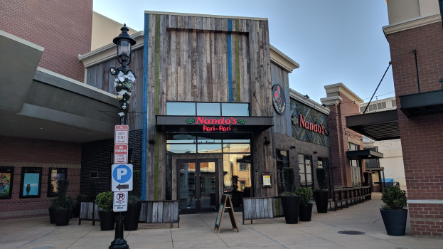 Außenansicht eines Restaurants mit einem Schild, umgeben von Gebäuden, Straßenmöbeln und Grünflächen unter einem klaren blauen Himmel.