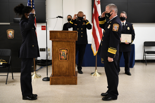 Eine Gruppe uniformierter Personen in Masken steht während einer Vereidigung eines neuen Feuerwehrchefs an einem Rednerpult und hält ein Zertifikat, mit Flaggen, Stühlen, Tischen und einer Wand im Hintergrund.