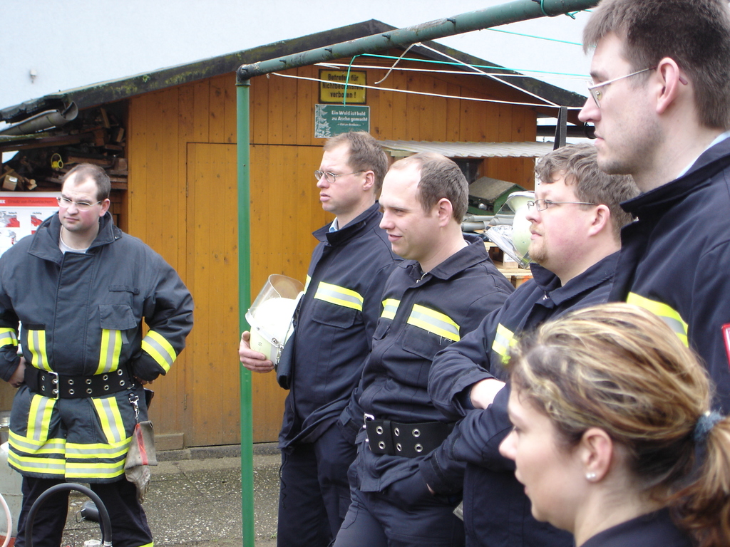 Eine Gruppe von Männern in blauen Feuerwehranzügen, die nach links schauen, mit einem kleinen Holzschuppen und Rohrrahmen im Hintergrund.