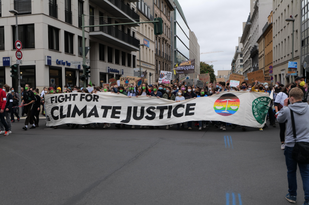 Eine Gruppe von Menschen marschiert mit einem Transparent, auf dem "Kampf für Klimagerechtigkeit" steht, eine Straße entlang. Im Hintergrund sind Gebäude, Schilder, Ampeln und Bäume zu sehen.