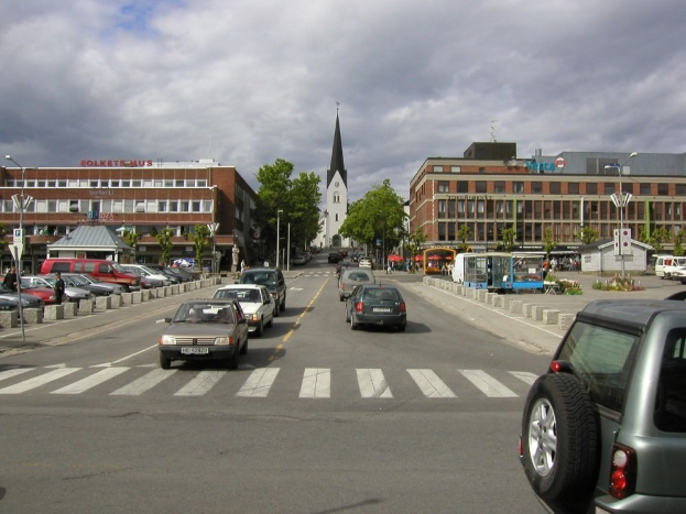 Stadtstraße mit parkenden Autos, Gebäuden, Bäumen, Laternen und einem bewölkten Himmel, mit einem sichtbaren Kfz-Kennzeichen.