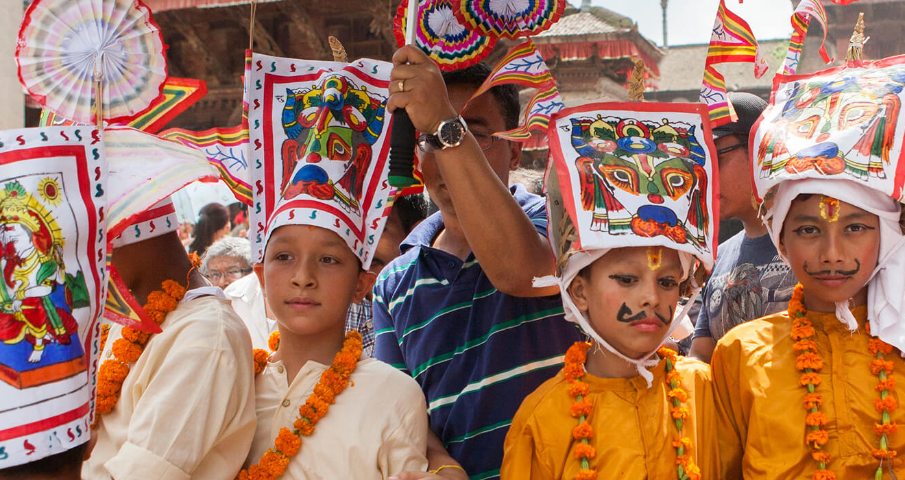 Eine Gruppe von Kindern in bunten Kostümen und Girlanden steht zusammen und hält Fahnen und Gegenstände, mit Gebäuden und Pfählen im Hintergrund unter einem klaren blauen Himmel, während eines Festivals in Kathmandu.