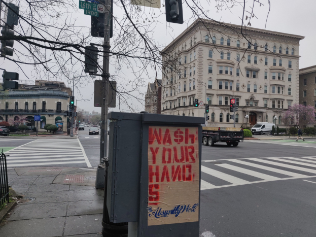 Eine belebte Stadtstraße mit Fahrzeugen, einem Fußgänger, einem "Waschen Sie Ihre Hände"-Müllcontainer, Verkehrsampeln, Schildern, einem Metallzaun, Grünflächen, Gebäuden und einem klaren blauen Himmel.