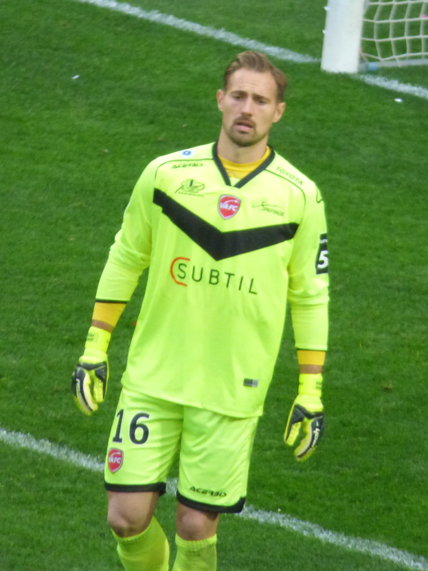 Ein Mann in einem gelben Torwarttrikot steht auf einem Fußballfeld mit einem Tor im Hintergrund, trägt ein T-Shirt, Shorts, Socken und Handschuhe.