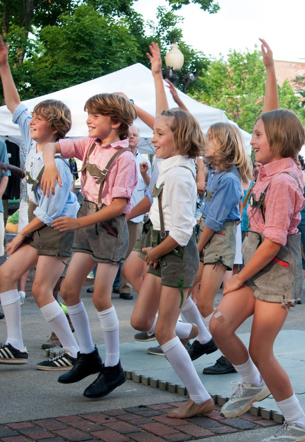 Kinder in traditioneller bayrischer Tracht tanzen auf einer Straße, umgeben von einer Menge, Zelten, Laternenmasten, Bäumen, Gebäuden und einem klaren blauen Himmel, einige tragen Shorts und Schuhe.