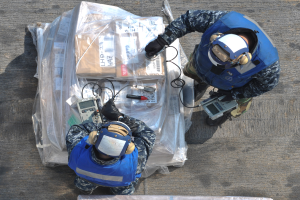 Zwei Männer in blauen Uniformen und Helmen arbeiten an einem großen Drohnenmodell, hältend verschiedene Gegenstände, mit einer Plastikabdeckung auf dem Boden vor ihnen.