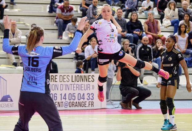 Eine Gruppe Frauen beim Handballspielen auf einem Court mit verstreuten Schuhen, während ein Publikum von Stühlen und Treppen aus zusieht und eine Person das Ereignis mit einer Kamera filmt.