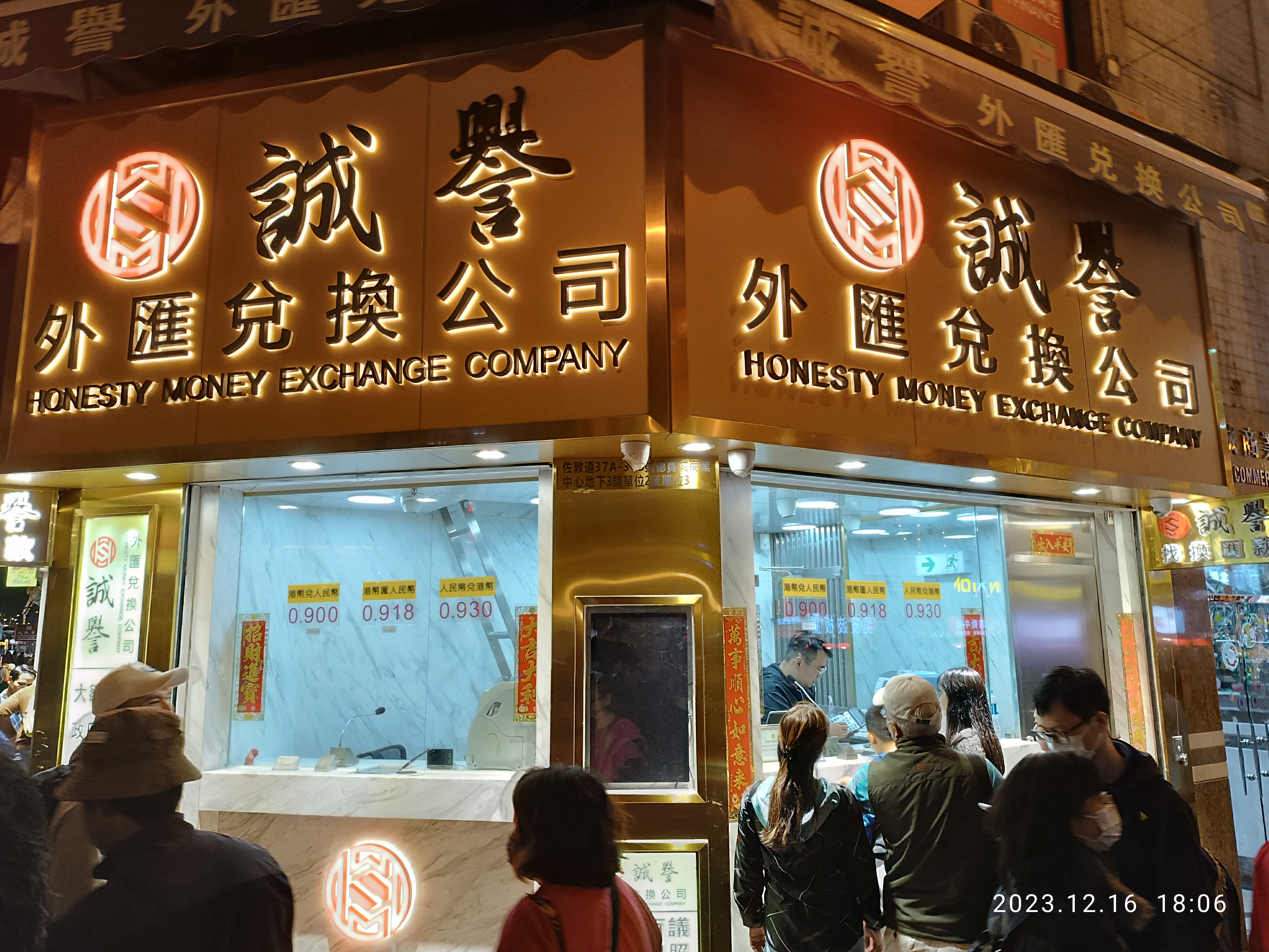 Eine Gruppe von Menschen steht nachts vor einem Gebäude, das von Lampen beleuchtet wird und von Schildern umgeben ist, einige tragen Taschen, mit einem Wasserzeichen im rechten unteren Eck, das auf eine ehrliche Geldwechsel-Firma hinweist.