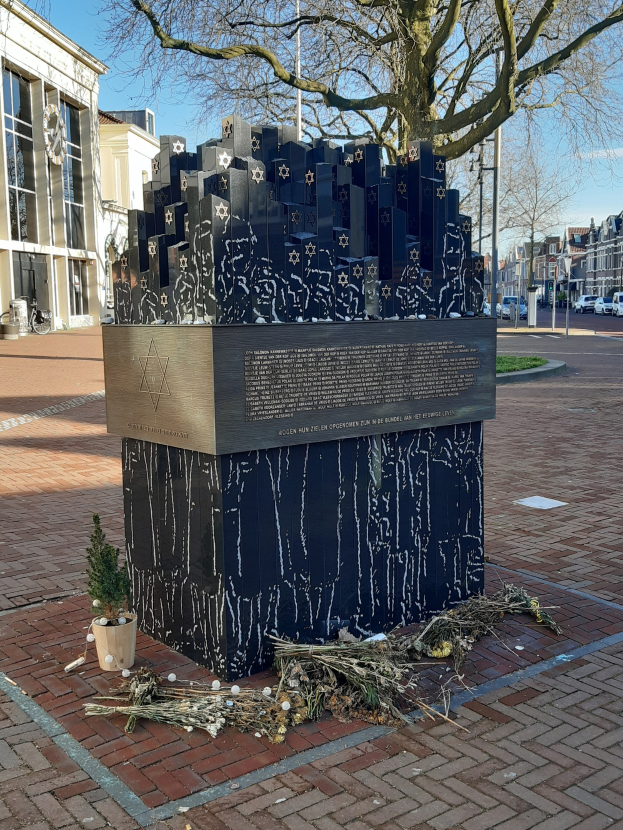 Denkmal in einem Stadtplatz für Holocaust-Opfer mit einem Baum davor, einem Topfgewächs auf dem Boden, verstreuten trockenen Blättern und Gebäuden, Fahrzeugen, Pfählen und Bäumen im Hintergrund.
