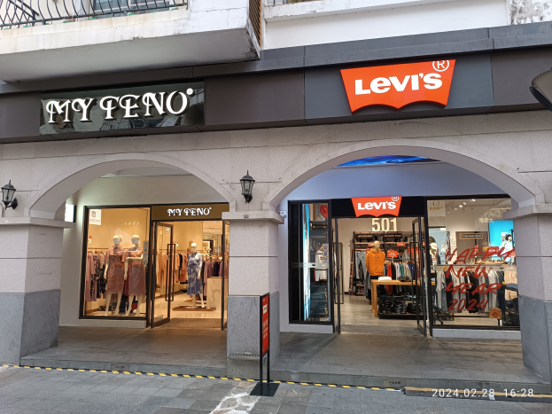 Storefront mit einem "Levi's"-Schild, Schaufensterpuppen in Kleidern, hängenden Kleidern, Beleuchtung, Texttafeln, Geländern und einer Straße unten.