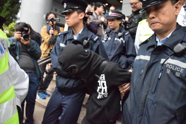 Eine Gruppe von Polizeibeamten steht vor einer Menge von Menschen, einige tragen Mützen und halten Kameras, mit einem Gebäude mit Säulen und Lampen im Hintergrund und Bäumen auf der linken Seite.