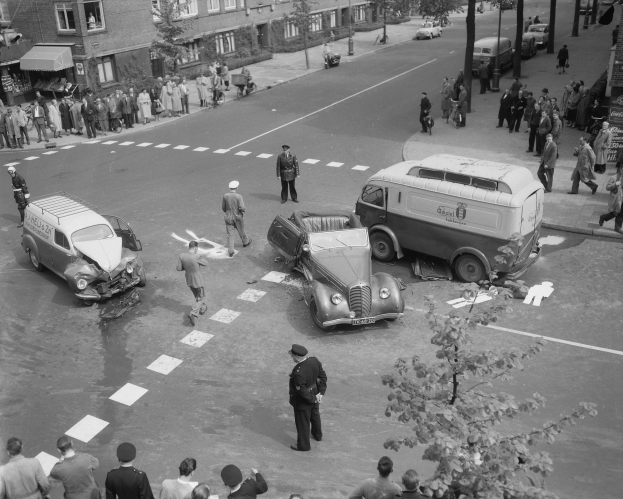 Schwarz-weiß-Szene eines Autounfalls an einer Straßenecke mit mehreren Fahrzeugen, darunter ein Van, und einer Gruppe von Menschen in der Nähe, mit Gebäuden, Bäumen, Pfählen und Texttafeln im Hintergrund.