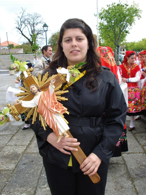 Eine Frau hält ein Abbild von Jesus Christus in ihrer Hand, mit ein paar anderen Menschen, die hinter ihr auf einem Bürgersteig stehen, und einem Hintergrund mit Bäumen, Laternenpfählen, Pflanzen, Straßen, Gebäuden und einem Metallpfahl.