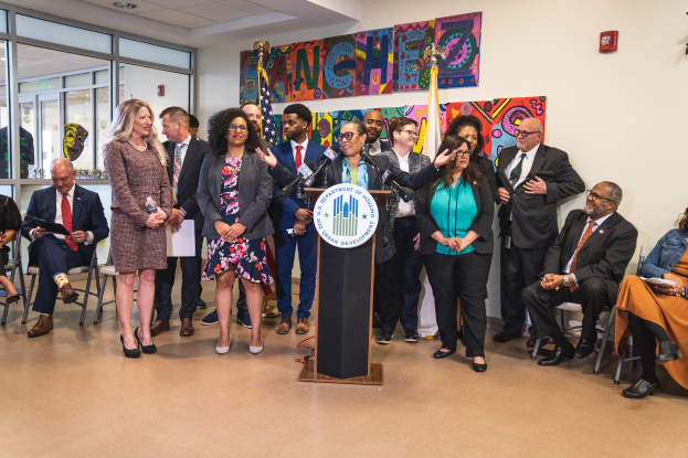 Gruppe von Menschen vor einem Rednerpult mit Mikrofon und einer Tafel mit der Aufschrift "Sancho Stadtratsmitglied tritt dem Stadtrat bei", mit sitzenden und stehenden Teilnehmern, Flaggen, Feuerlöscher und Deckenleuchten im Hintergrund.
