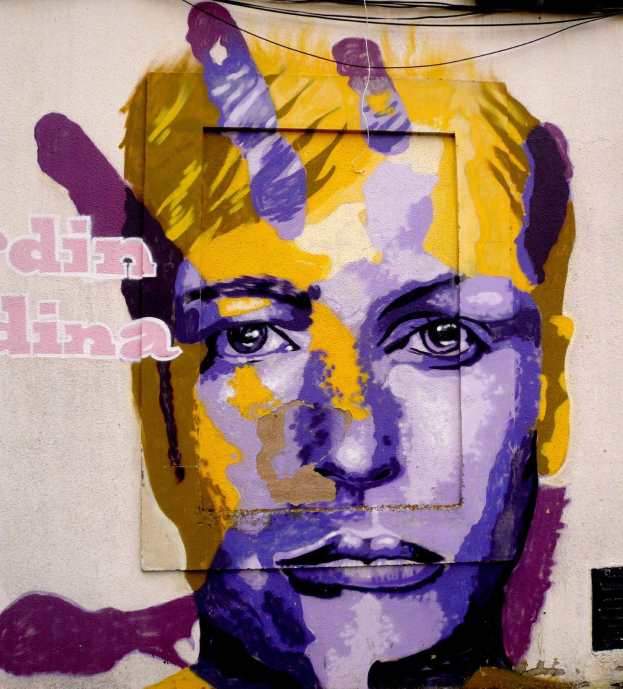 Ein farbenfrohes Wandgemälde eines Frauenkopfs auf einem Gebäude in Berlin mit detaillierten Zügen, direktem Blick und blaugrün-gelben Buchstaben.