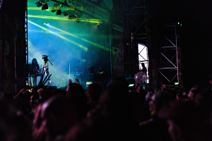 Ein volles Konzert mit Musikern auf der Bühne, die Instrumente spielen, unter Scheinwerfern, mit einem Banner, Metallrahmen und Mikrofonständern im Bild.