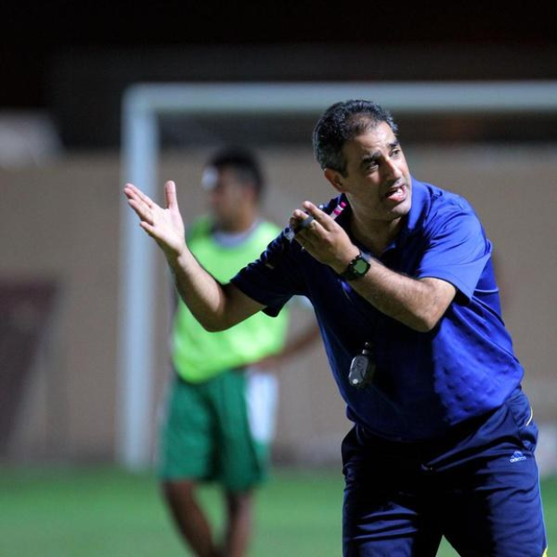 Ein Mann in einem blauen T-Shirt und Hose steht auf einem Fußballfeld und hält ein Mikrofon in der Hand, während er als Trainer gestikuliert. Im Hintergrund sind eine Person in einem grünen Kleid, ein Torpfosten und eine Wand zu sehen.