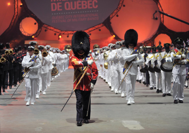 Eine Gruppe uniformierter Personen marschiert auf einer Straße, einige spielen Musikinstrumente, mit einem beleuchteten Schild im Hintergrund, das die Eröffnungszeremonie des Montreal International Festivals of Military Bands anzeigt.