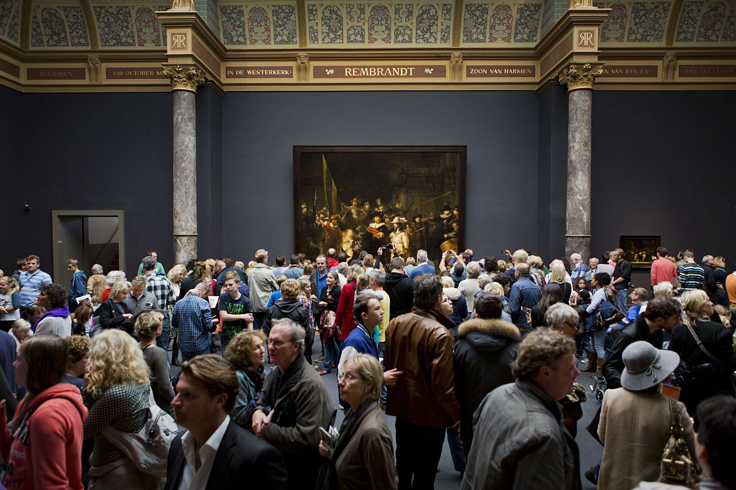 Eine gro├če Gruppe von Menschen in einem Raum mit einem Foto auf der Wand, Pfeilern, einer T├╝r und einer Decke mit Lichtern, einige halten Taschen, im Museum of Fine Arts in Amsterdam.