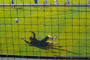 Ein Fussballer hält den Ball vor einem Tor, mit einem Netz im Vordergrund und einigen Menschen im Hintergrund, sowie mehreren Bällen auf dem Boden verstreut.