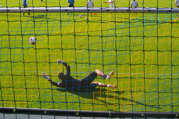 Ein Fussballer hält den Ball vor einem Tor, mit einem Netz im Vordergrund und einigen Menschen im Hintergrund, sowie mehreren Bällen auf dem Boden verstreut.