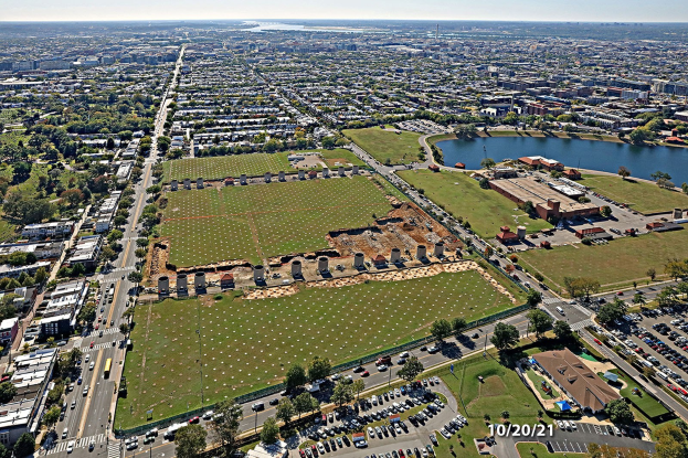 Ein Park mit grünem Gras, Bäumen, Pflanzen, Laternenmasten und Skulpturen, mit Gebäuden und einem klaren blauen Himmel im Hintergrund.