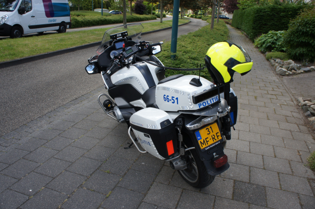 Ein Polizeimotorrad steht am Straßenrand mit einem Helm darauf, umgeben von Bäumen, Pfählen, Gebäuden, Pflanzen, Gras, Steinen und anderen Fahrzeugen.
