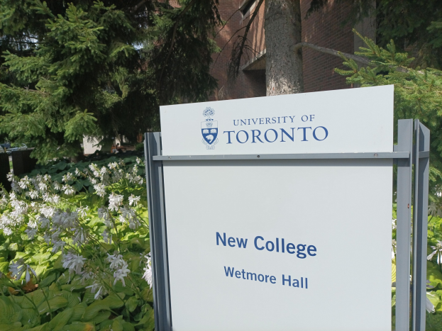 Außenansicht des neuen Colleges der Universität Toronto an Wetmore Hall, mit einem Schild vor dem Gebäude umgeben von grünem Gras, bunten Blumen, hohen Bäumen und einem klaren blauen Himmel.