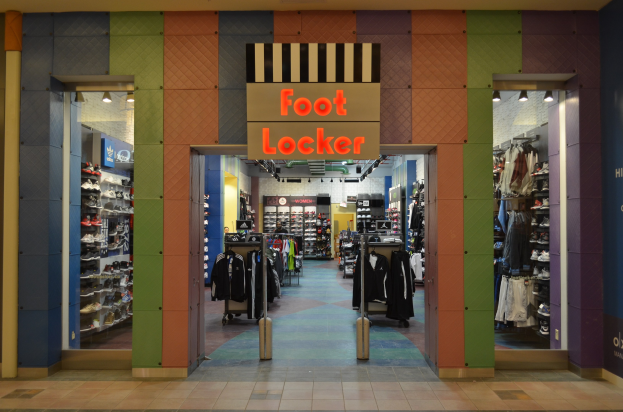 Innenansicht eines Foot Locker-Schuhgeschäfts mit Kleidung an Bügeln, Waren auf Gestellen und Deckenbeleuchtung, die den Boden zeigt.