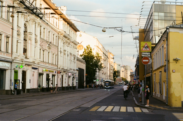 Stadtstraße mit Fußgängern und Radfahrern, gesäumt von Gebäuden, Schildern, Bäumen und Strommasten.